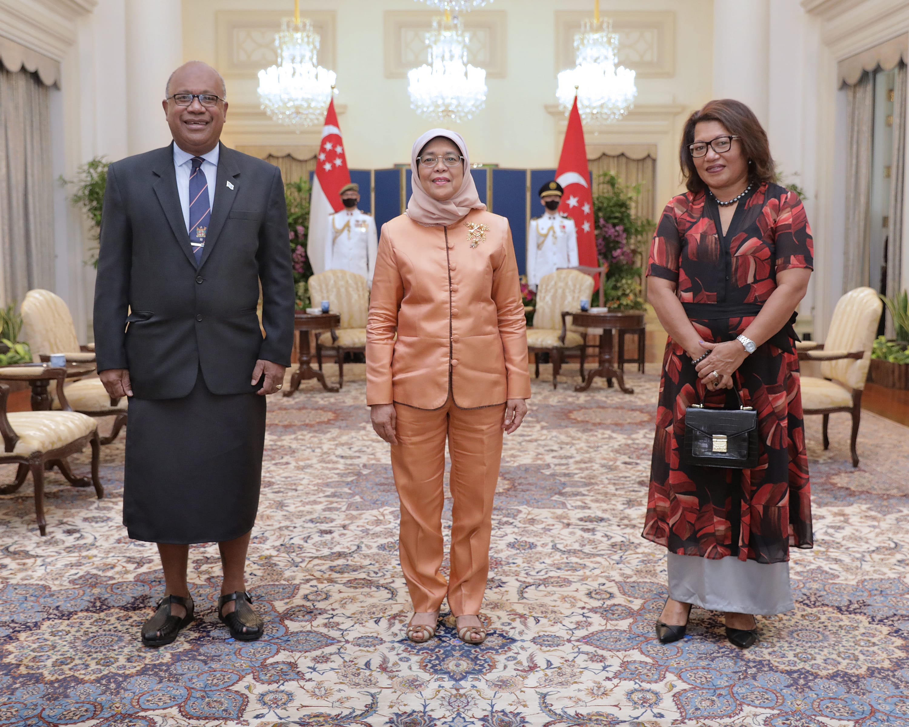 Three people stand indoors with Singapore flags behind them. One wears a hijab and peach pantsuit.